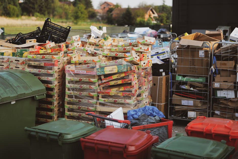 An outdoor scene depicting a large collection of waste materials and recycled goods arranged on a paved surface, likely in a private property or a waste management area. The foreground features severa
