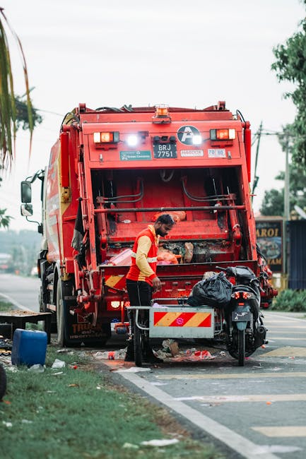 An outdoor scene showing a large collection of overflowing rubbish bins and scattered waste materials on a paved area near a parking lot. The waste includes cardboard boxes, plastic bags filled with r