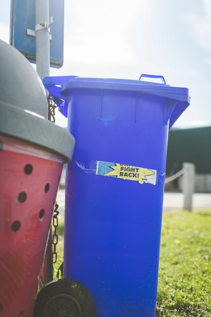 A large green wheeled waste bin filled with multiple red and black plastic bags containing unknown rubbish, placed on a paved surface adjacent to a dark brick wall. The bin appears to be used for priv